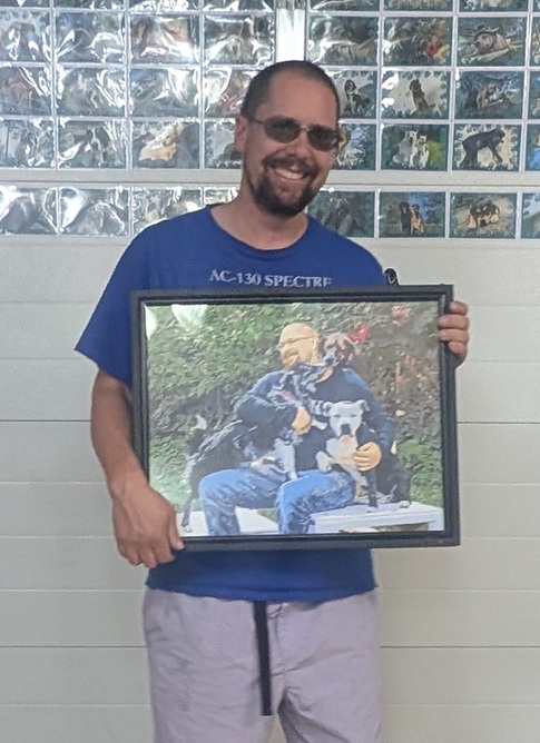 Dog trainer standing in front of a wall of dog pictures holding his completed painting of him hugging three dogs.