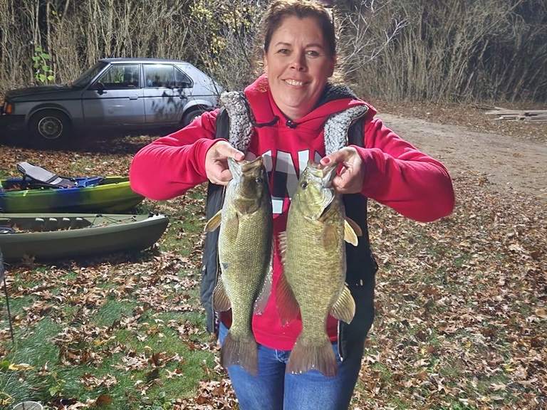 A woman in a red sweatshirt is holding up two sizeable smallmouth bass outside in a fall setting.