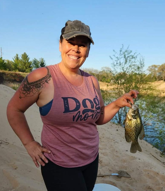 A smiling woman in a pink t-shirt that says "Dog mom" is holding up a small fish and laughing on a sand beach in front of a lake.